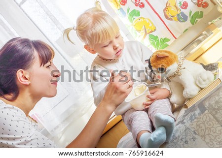 Mother feeding baby food. Young woman feeding kid daughter yogurt on kitchen. Little girl eating yogurt.