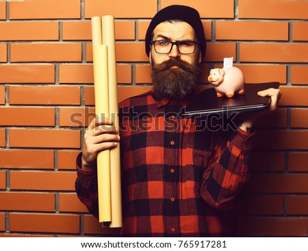 Bearded man, long beard. Brutal caucasian serious unshaven hipster holding craft paper rolls, piggy bank on laptop in checkered shirt with hat and glasses on brown brick wall studio background