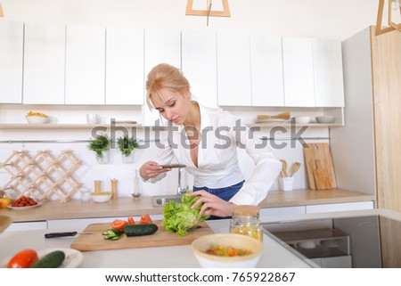 Nice young woman takes photo on smart phone and puts in frame leaves of green salad and cucumber with red tomato on cutting board with knife, smiles and forms composition for photo in blog or social