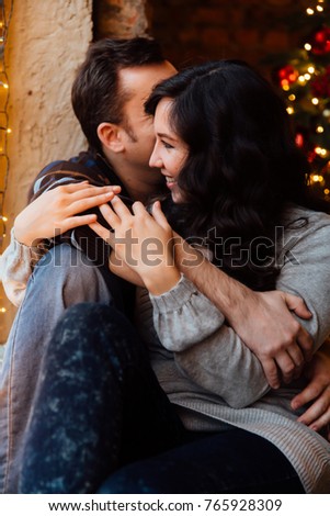 Couple of lovers hugs sitting on the windowsill in the Christmas loft studio. guy is hugging the girl. Garlands and New Year's lights. backdrop of a Christmas tree.