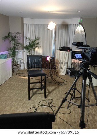 A small room setup for an interview style video shoot behind the scenes. Motion picture camera, lighting, reflector lavaliere microphone setup with plants in background for portrait framed person