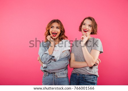 Picture of cheerful two women friends standing isolated over pink background. Looking aside.