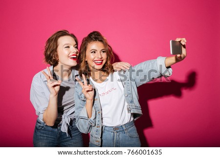 Image of cheerful two women friends standing isolated over pink background. Looking aside make selfie by phone showing peace gesture.