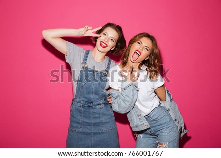 Photo of emotional two women friends standing isolated over pink background. Looking camera showing peace and rock gesture.