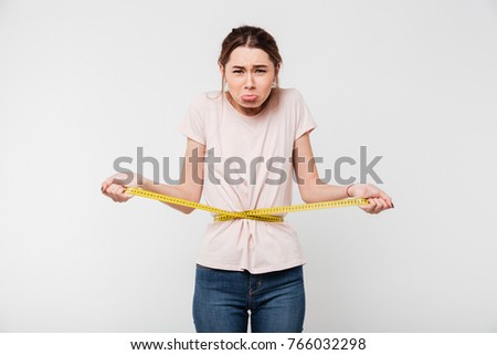 Portrait of an upset sad woman holding measuring tape around her waist and looking at camera isolated over white background