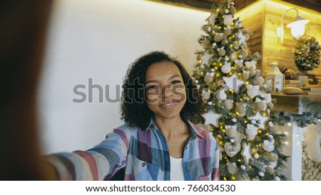 Funny mixed race girl taking selfie pictures on smartphone camera at home near Christmas tree