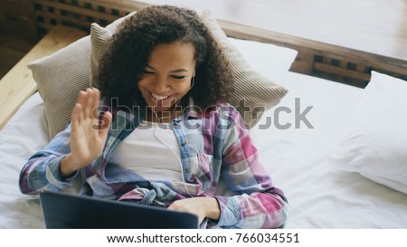 Cheerful mixed race girl having video chat with friends using laptop camera while lying on bed Royalty-Free Stock Photo #766034551