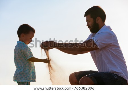 Happy family on sunset sea beach. Father, baby son play with sand slipping through man hand fingers. Child catching falling sand. Active parents, people outdoor activity on summer vacations with kids.