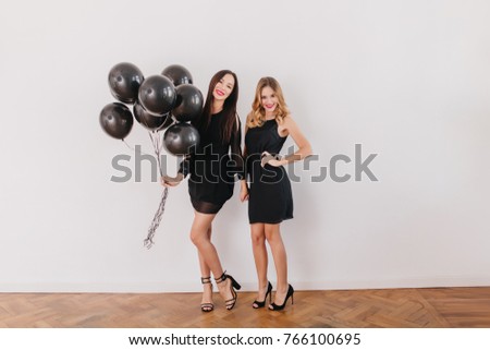 Full-length portrait of slim caucasian girls in black dresses preparing for birthday party. Graceful brunette lady wears sandals holding helium balloons while posing with sister in her room.