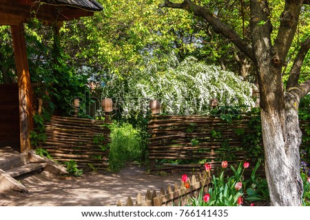 A traditional Ukrainian courtyard in the spring with an outlet to the garden and garden, with flowers in a flowerbed, a wattle fence decorated with clay pots, ivy and fruit trees. Royalty-Free Stock Photo #766141435