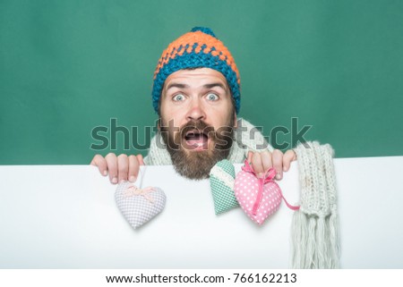 Holiday celebration and party. Valentines day and love. Hipster with heart on green white background. Guy with surprised face in winter hat and scarf. Man with long beard hold heart pillow.