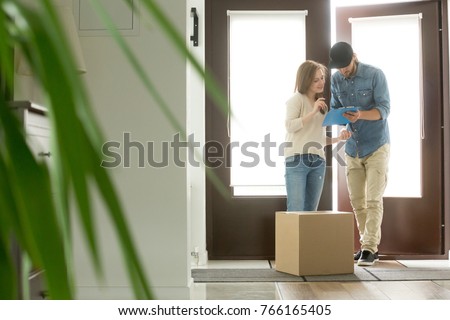 Young woman receiving cardboard box from courier at home, receiver about to sign document for accepting parcel, man with clipboard delivering package to customer, door delivery service concept Royalty-Free Stock Photo #766165405