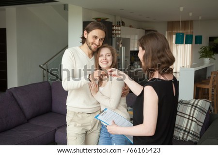Happy young couple becoming tenants getting own keys of new home at meeting with real estate agent indoors, female realtor holding rental agreement making house for rent deal with satisfied clients