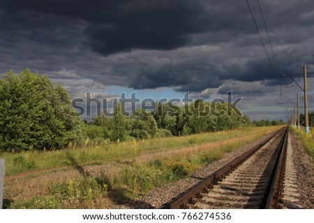 Railroad receding into the distance beyond the horizon on the backdrop of an impending storm clouds in the center of which a clear blue sky to the left of the road green grass and trees