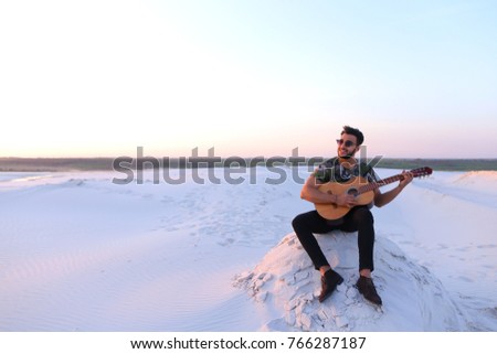 Handsome guy, Arab playing with pleasure on musical stringed instrument and with beautiful smile on face. Man sings and drives guitar strings, sitting on hill in middle of wide sandy desert on warm