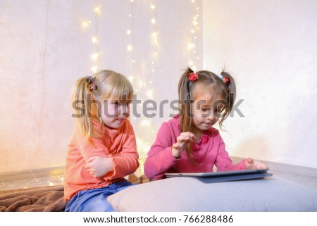 Cute little girls play in computer games on device or watch cartoons, press something on touch screen of tablet, smile and communicate with each other, sitting on rug in bright room with flickering