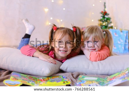 Funny little girls friends read children's bright book and look at pictures, laugh and smile at each other, play games and talk, pose on camera and lie on soft cushions in bright room with New Year's