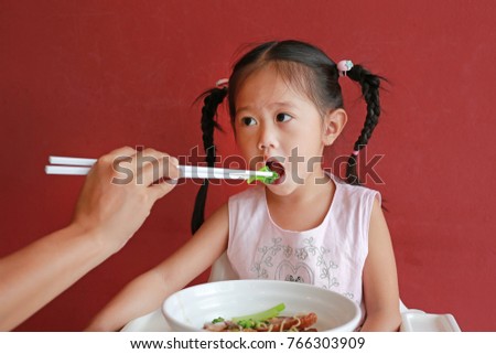 Mother feeding vegetable for her daughter on high chair against red wall background.