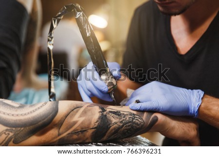 Close up of professional tattooer artist doing picture on hand of man by machine black ink from a jar. Tattoo art on body. Equipment for making tattoo art. Master makes tattooed in light studio