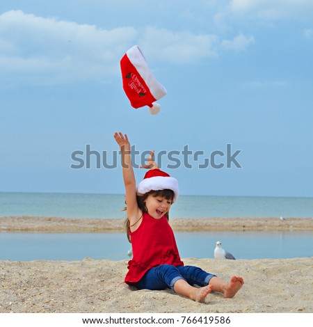 Cute little girl celebrating Christmas and New Year holidays on the beach, throwing up Santa hat