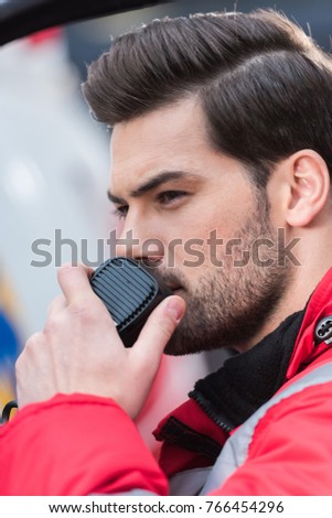 portrait of young handsome male paramedic standing near ambulance and talking by portable radio