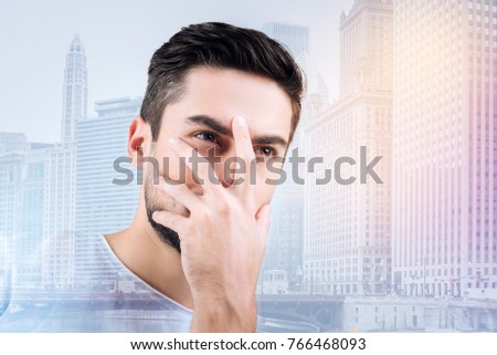 Little bit shy. Concentrated male person raising left hand while covering his face, standing against urban surroundings