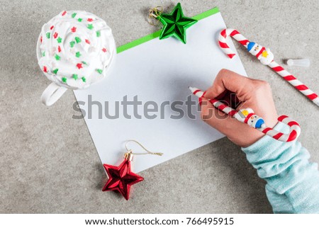 A gray table with a greeting sheet, Christmas decorations, a cup of hot chocolate and pen in form of candy cane. Girl writing, female hand in picture, copy space top view