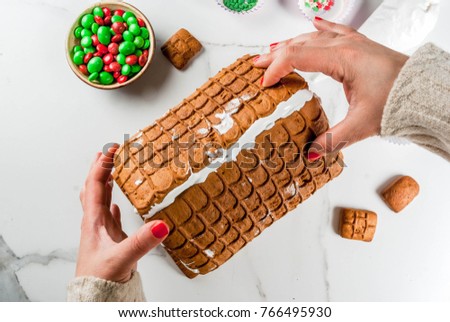 Preparation for Christmas, New Year. Cooking and decoration of traditional advent gingerbread house, female hands in picture, top view, white marble background.