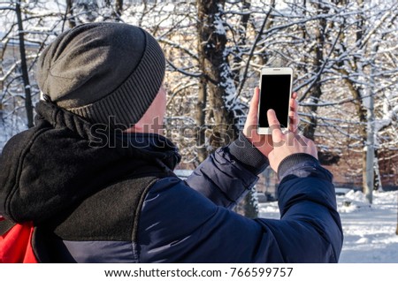 Trendy young man standing and using white mobile smartphone with a red backpack in the snow-covered pine forest on a cold winter day