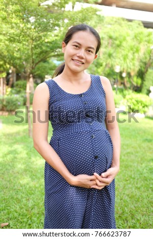 Pregnant woman standing in the green summer garden with looking at camera.