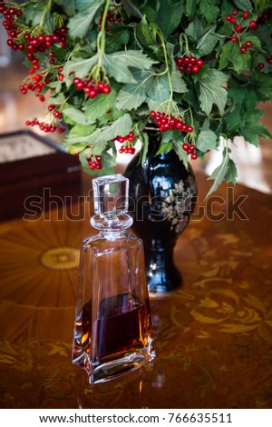 A bottle of alcohol stands on a wooden table on a blurred background. Bottle with cognac. Bottle with cognac next to the vase. Red viburnum stands in a vase next to a bottle of alcohol.