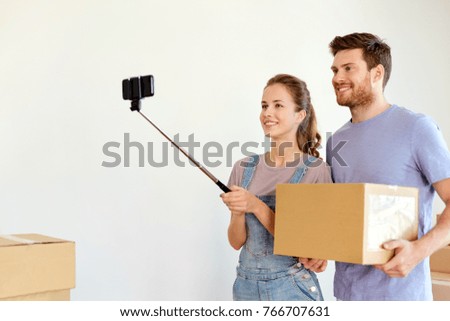 moving, technology and real estate concept - happy couple with boxes and smartphone on selfie stick taking picture at new home