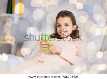 people, children and technology concept - happy smiling girl lying awake with smartphone in bed over holidays lights background