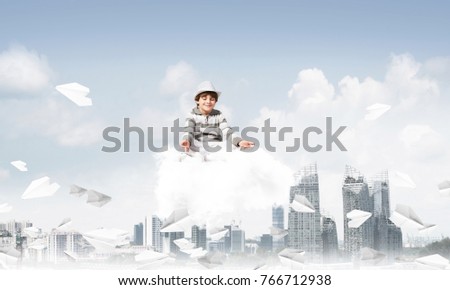 Young little boy keeping eyes closed and looking concentrated while meditating on cloud among flying paper planes with cityscape view on background.