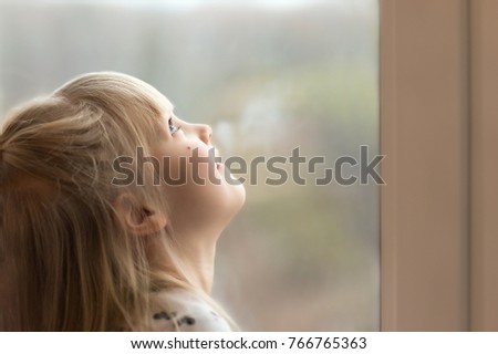 Little cute blond girl dreaming and looking outside window, waiting for something.