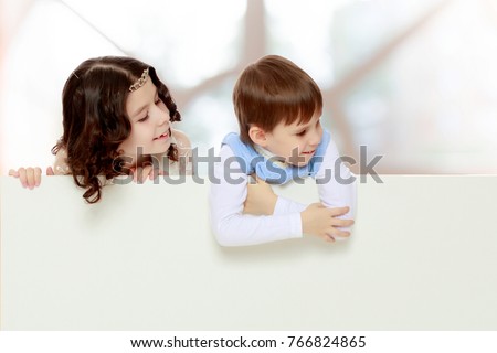 A boy and a girl peeping from behind the white banner.Children look to the side.In a room with a large semi-circular window.