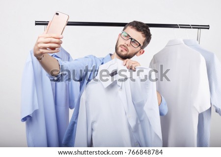 Metrosexual Caucasian guy taking self portrait on generic mobile phone, posing against blue background of his wardrobe, boasting about new stylish jeans and shirts she bought in the sale this morning.
