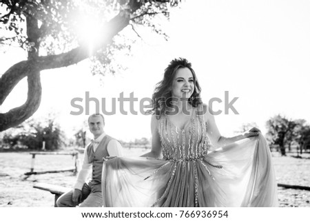 Wedding at sunset. Couple is sitting on a bench under a tree. Beige dress with sparkles. Light suit with a bow tie. The bride and groom embrace and kiss. Black and white photo