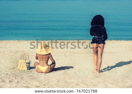 Female in a yellow swimsuit and hat and a girl in a fur coat on the beach. Conceptual picture of winter and summer. Winter is leaving. Summer is leaving. Two seasons winter and summer.