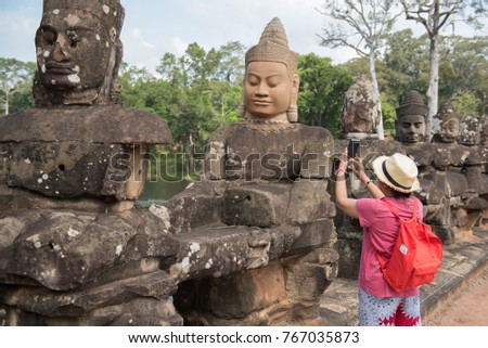 Young female with red backpack using mobile phone or cell phone camera taking a photo of statues at South Gate Angkor Thom Temple complex, Angkor Wat, Siem Reap, Cambodia, traveling concept.