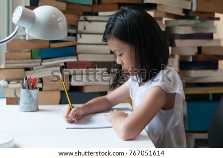 Young Asian teenage girl writinng in note book with blurred book pile and pencil cup in background. Education theme
