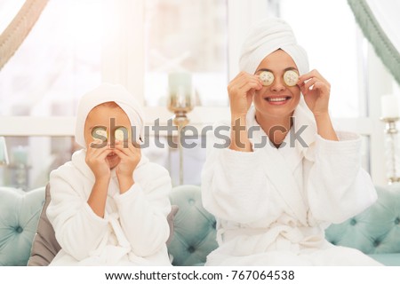 Photo of mother and daughter in white bathrobes. They are sitting on the couch and applying pieces of cucumber to their eyes. Their hair is wrapped in a white bath towel. Royalty-Free Stock Photo #767064538