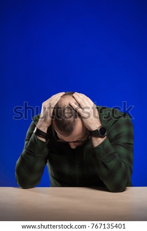 Emotional man in glasses sits at a table and posing on a blue background