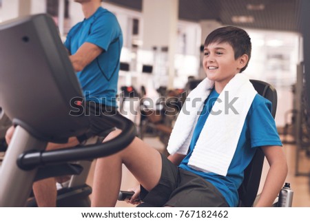 Father with his son in the same clothes in the gym. Father and son spend time together and lead a healthy lifestyle. The boy is engaged in simulators, the trainer is next to him.