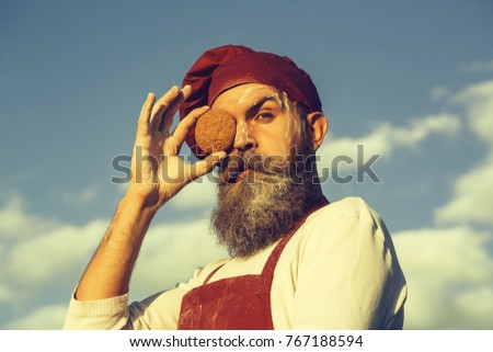 Handsome bearded man cook chef in uniform and red hat and apron with long beard holding oatmeal cookies near serious face sunny day outdoor on blue sky background