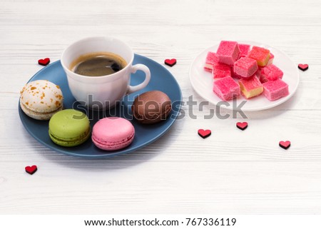 A cup of coffee with french colorful macaroons, marmalade on a white wooden table, decorated with hearts. St. Valentine's Day.