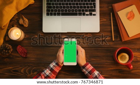Top view of woman hands holding white smartphone with green screen, tapping, scrolling with her finger. Autumn top view. Chroma key