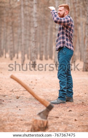 A man is standing looking into the distance holding his hand to his forehead. Near the stump with an ax. Outdoors.