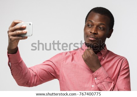 Horizontal portrait of handsome African American guy isolated on white background dressed in casual shirt looking content and happy while taking selfie with phone he is holding in stretched arm