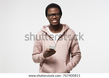 Closeup portrait of young African American man in casual clothes and spectacles pictured isolated on white background holding cellphone in hand looking at camera with open smile, browsing or texting